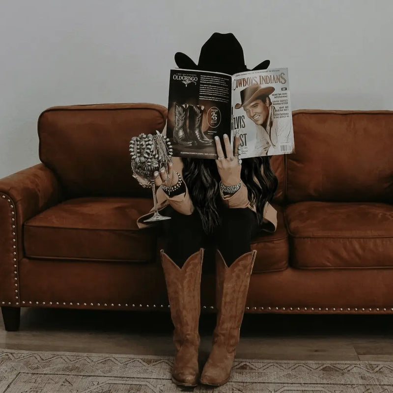 A person in a cowboy hat and boots sits on a brown couch, holding a magazine and a bedazzled wine glass. Their face is hidden behind the magazine.