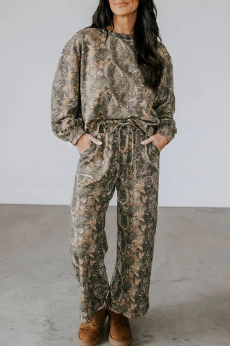 A woman stands indoors wearing a loose-fitting, long-sleeve camouflage jumpsuit with pockets and brown shoes. Her hands are in her pockets, and her long dark hair is down.