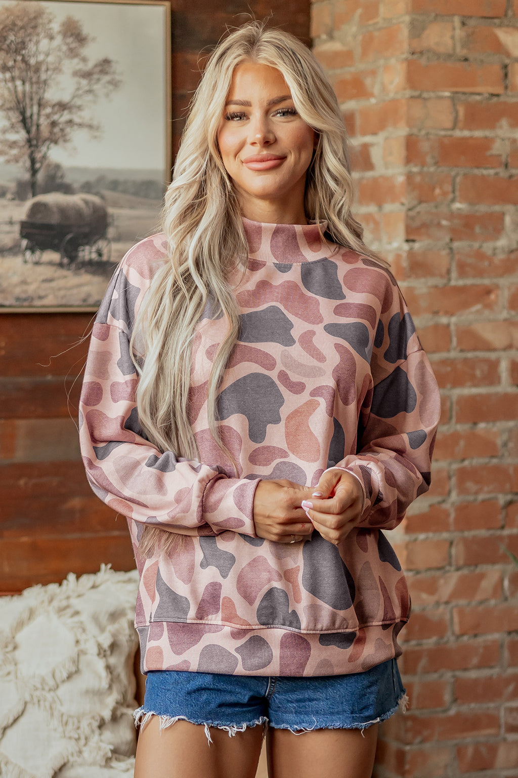 A woman with long blonde hair, wearing the Camo Mock Neck Pullover Sweatshirt in pink and purple with denim shorts, stands in front of a brick wall, smiling softly.