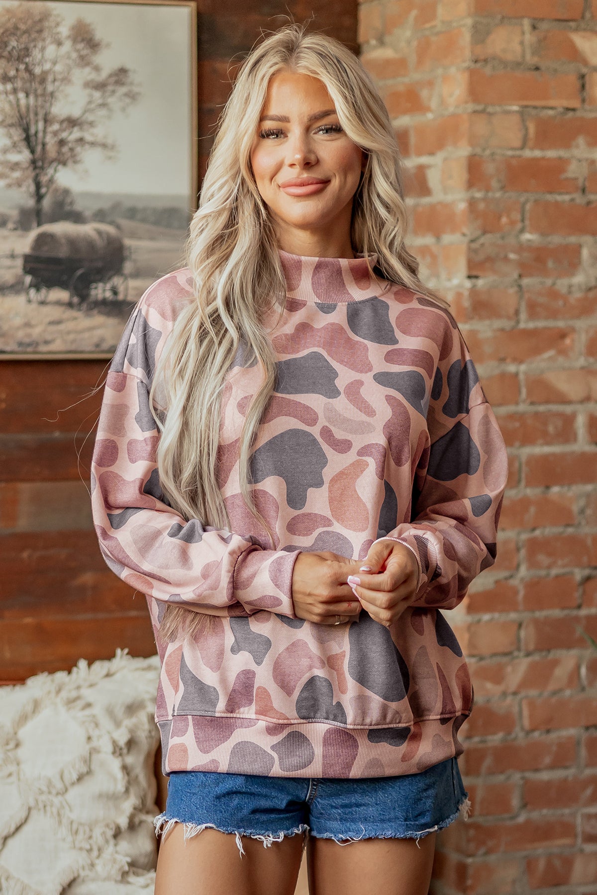 A woman with long blonde hair, wearing the Camo Mock Neck Pullover Sweatshirt in pink and purple with denim shorts, stands in front of a brick wall, smiling softly.