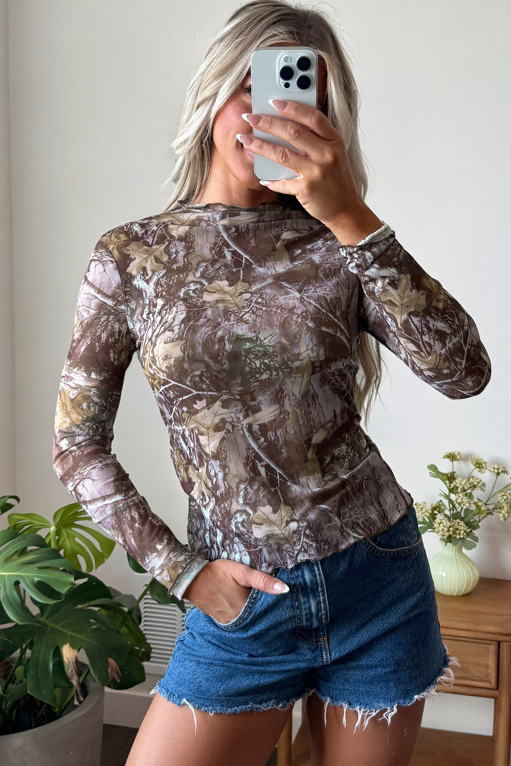 A person with long, light hair takes a mirror selfie wearing the Smoke Gray Floral Mesh Mock Neck Top and frayed denim shorts, creating a slim silhouette. There are potted plants and a small vase with flowers in the background.