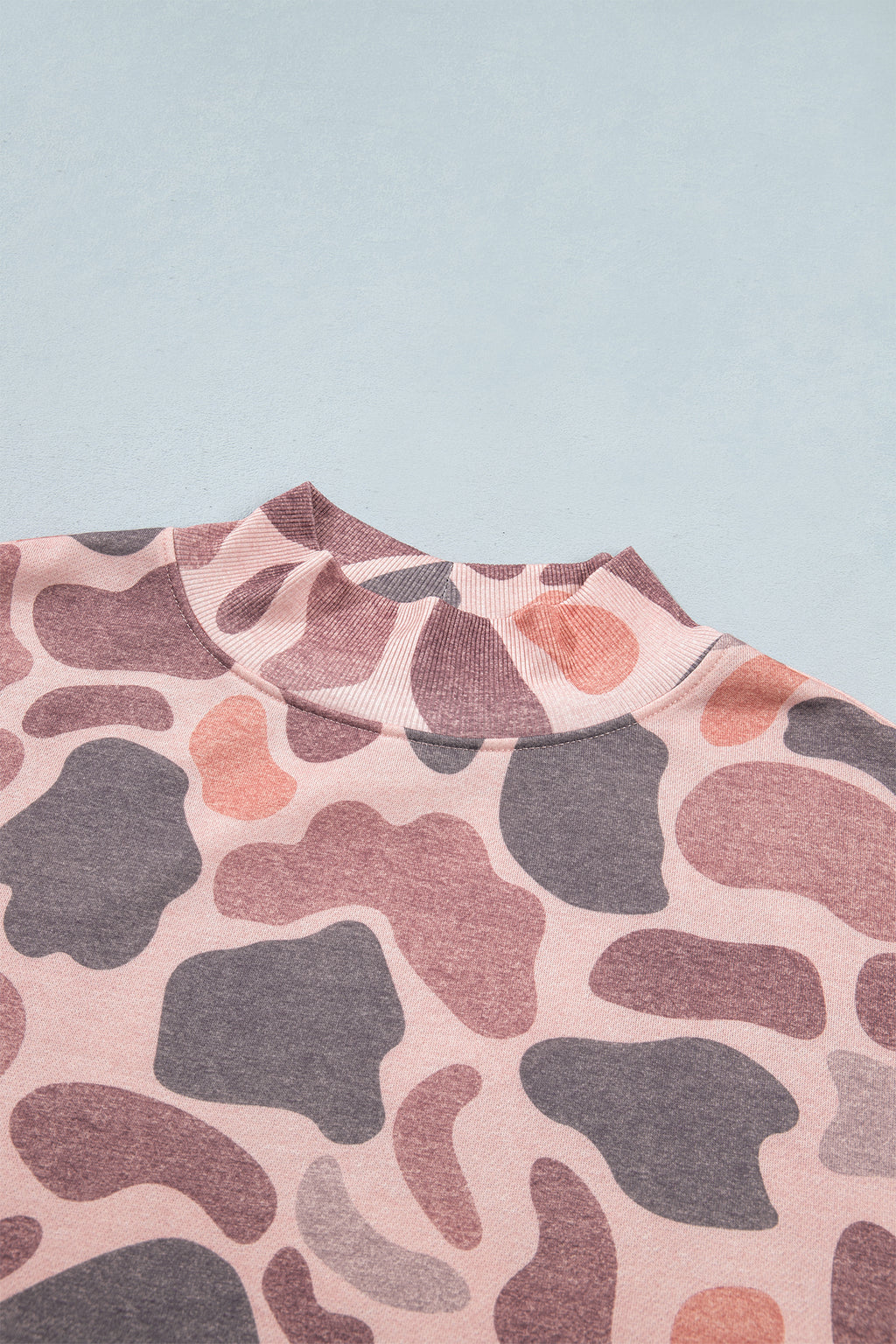 A close-up of the Camo Mock Neck Pullover Sweatshirt in pink and mauve abstract camo print, featuring a ribbed mock neckline and laid flat on a light blue surface, highlights its unique style and everyday comfort.