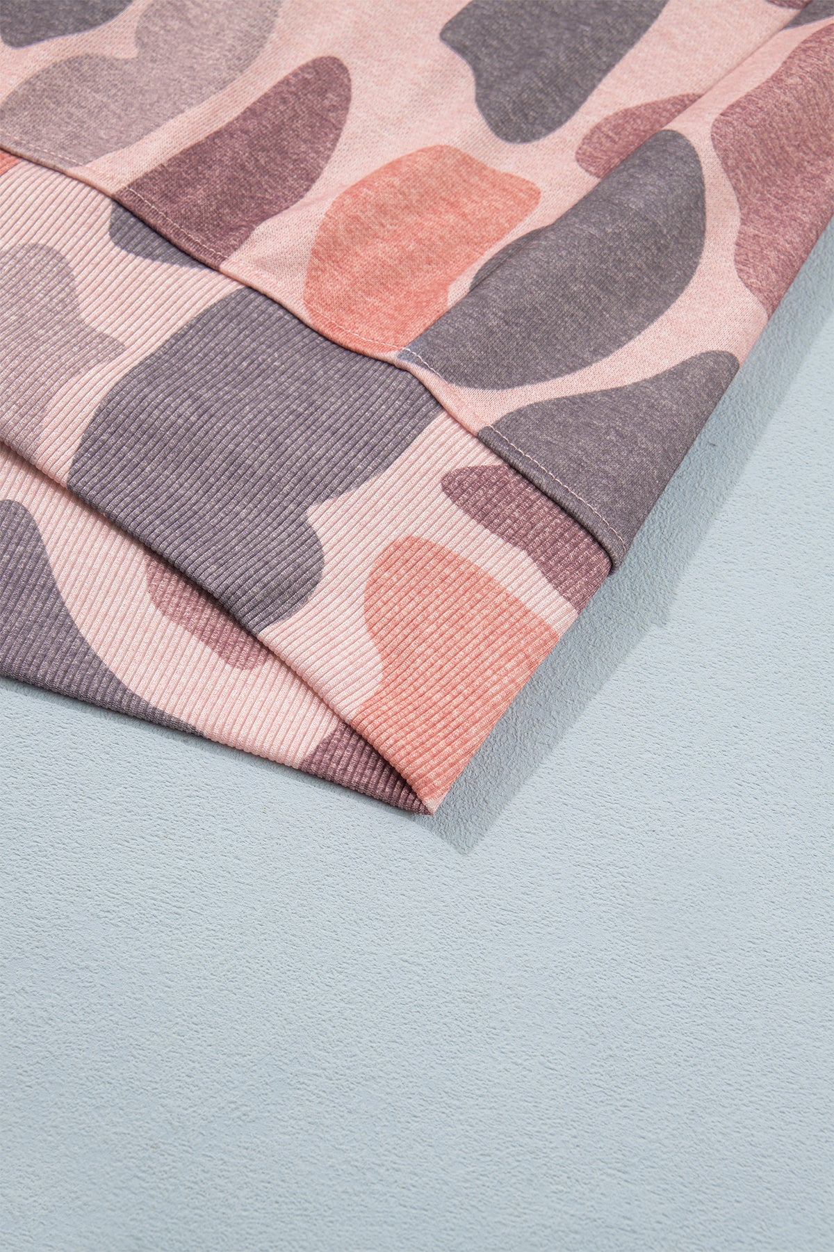 A folded Camo Mock Neck Pullover Sweatshirt featuring a pink, grey, and mauve abstract pattern rests on a light blue surface. Reminiscent of western-inspired camo styles, the sweatshirt’s textured fabric and overlapping layers are clearly visible.