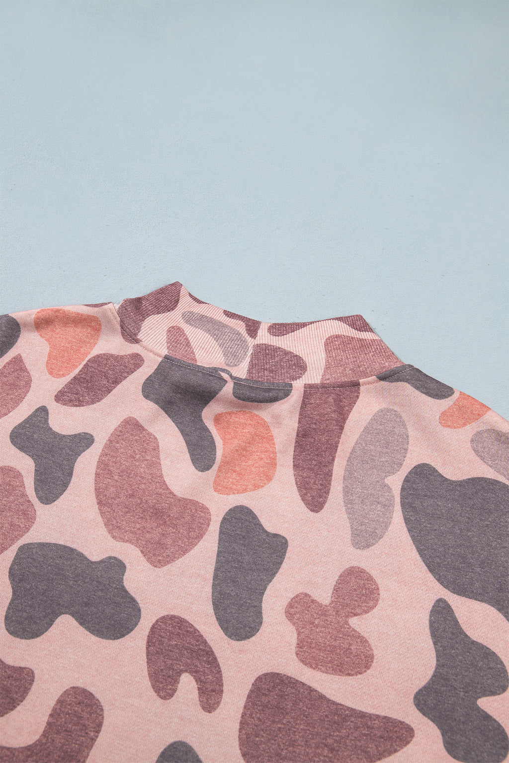 A close-up of the Camo Mock Neck Pullover Sweatshirt, featuring a pink, purple, and coral camouflage pattern, lies flat on a light blue surface, highlighting the collar and upper back.