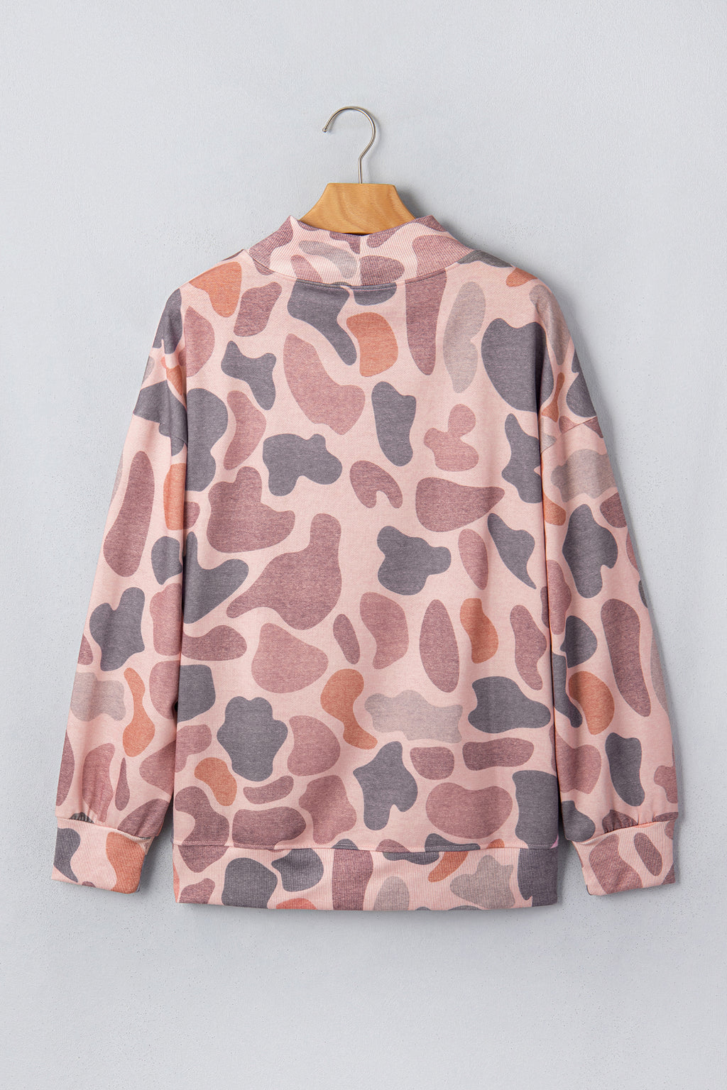 The Camo Mock Neck Pullover Sweatshirt, featuring a long-sleeve pink, gray, and brown camouflage pattern, hangs on a wooden hanger against a light gray wall.