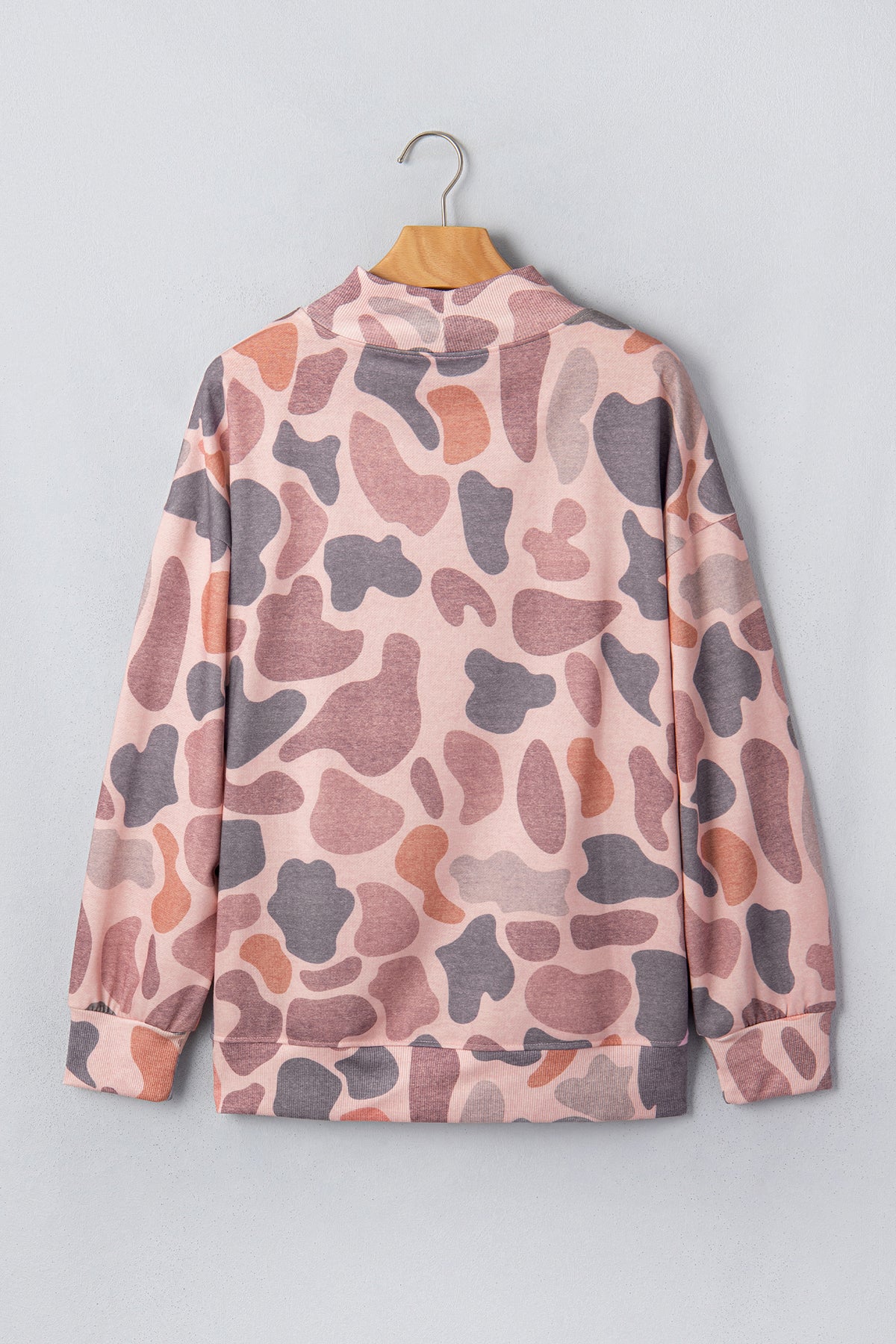 The Camo Mock Neck Pullover Sweatshirt, featuring a long-sleeve pink, gray, and brown camouflage pattern, hangs on a wooden hanger against a light gray wall.
