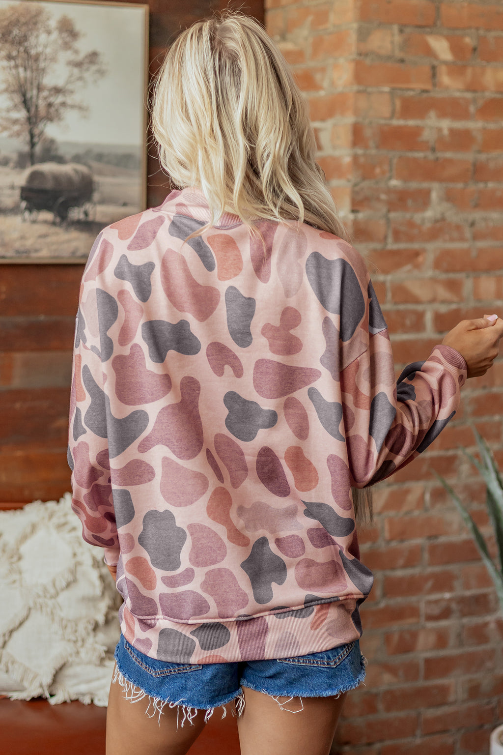 A person with long blonde hair wears the Camo Mock Neck Pullover Sweatshirt in pink and gray, paired with denim shorts, standing with their back to the camera in a cozy room featuring a brick wall and a framed picture.