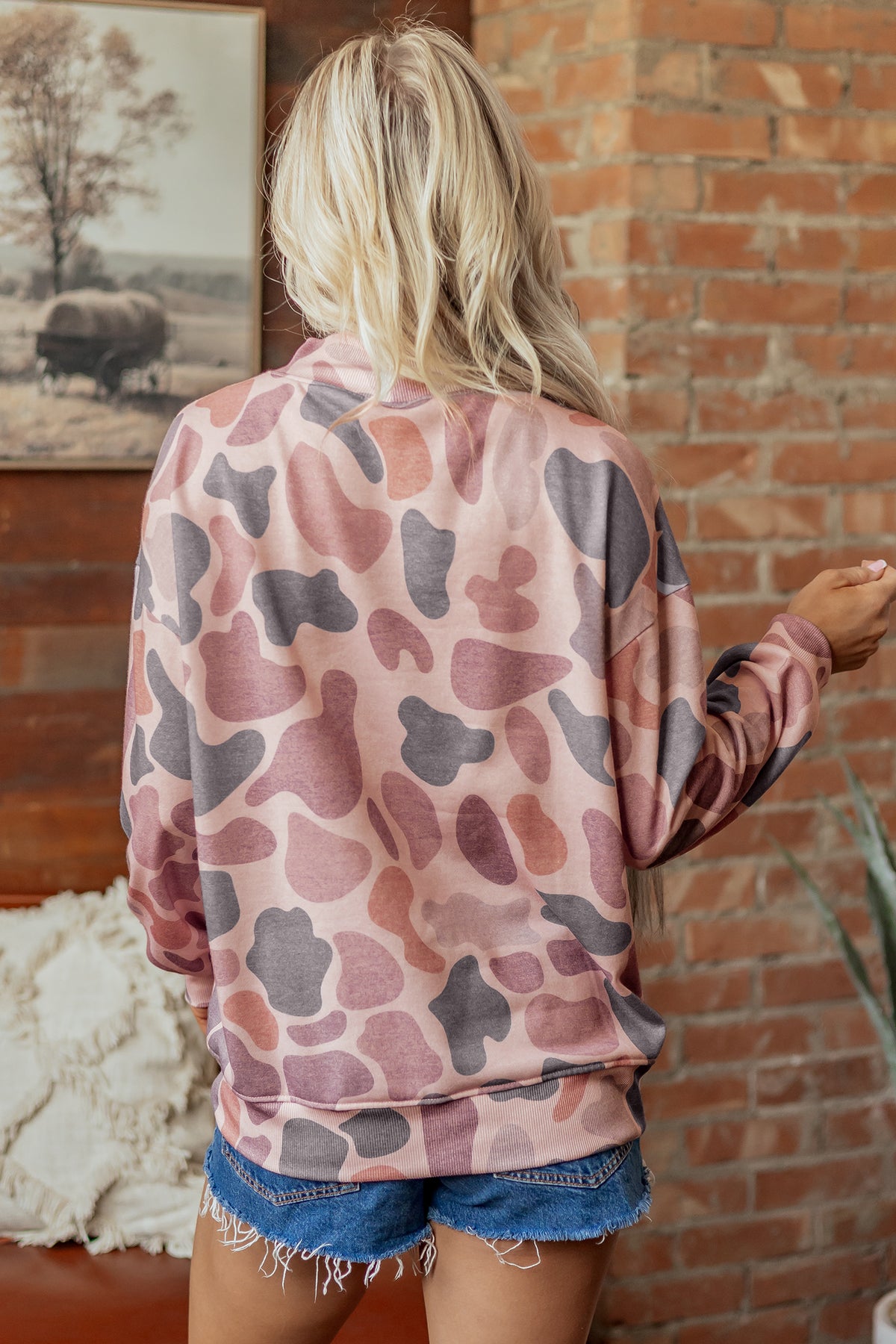 A person with long blonde hair wears the Camo Mock Neck Pullover Sweatshirt in pink and gray, paired with denim shorts, standing with their back to the camera in a cozy room featuring a brick wall and a framed picture.