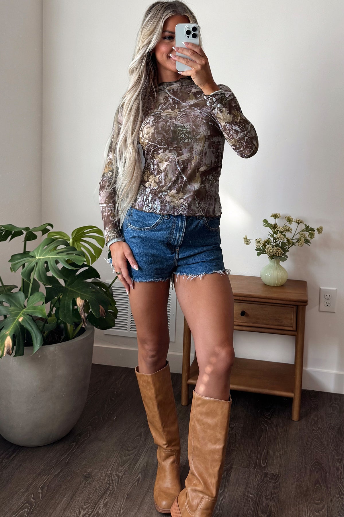 Woman taking a mirror selfie, wearing the Smoke Gray Floral Mesh Mock Neck Top paired with denim shorts and tan knee-high boots. She stands by a wooden table with a small plant on it and a large potted plant on the floor.