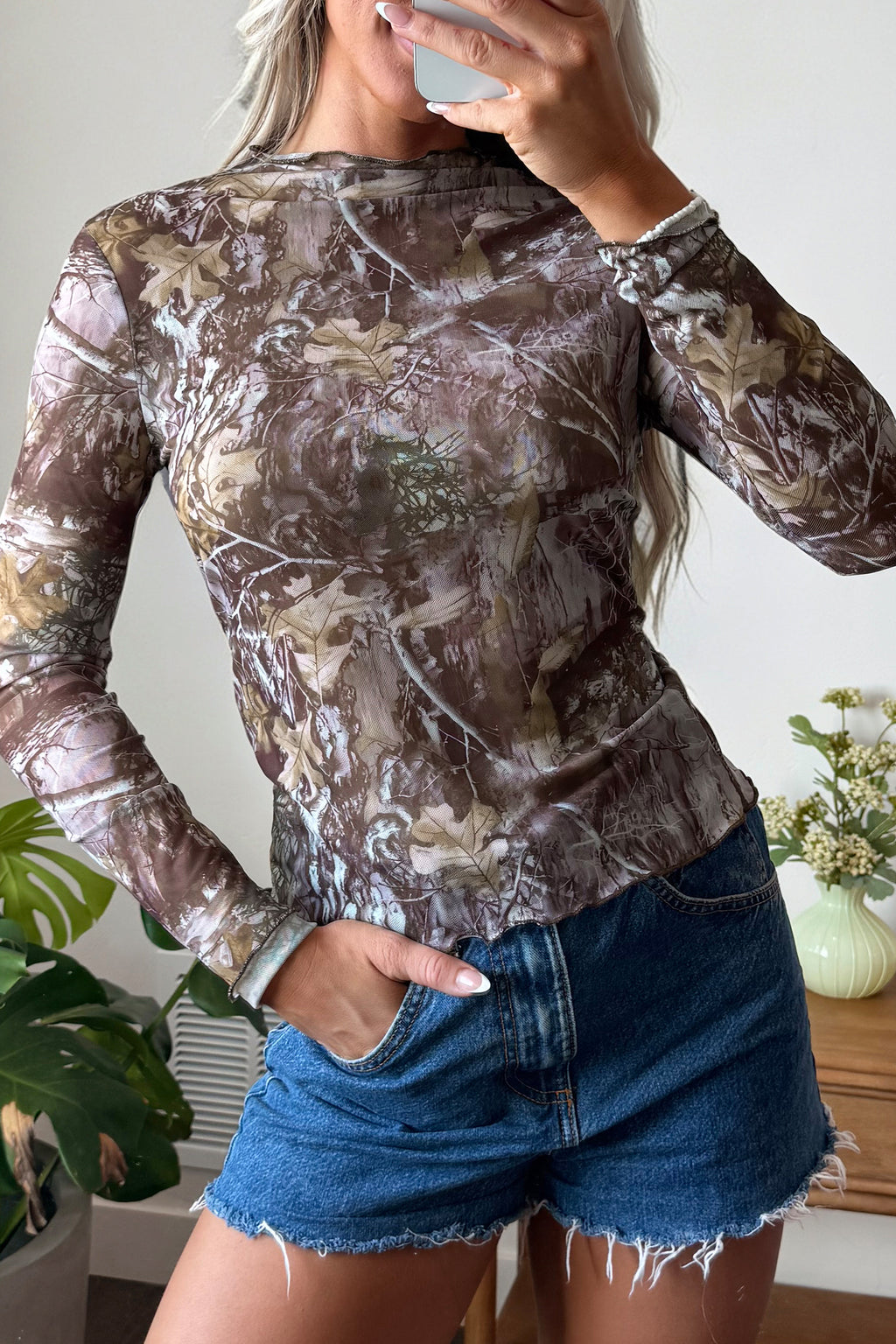A person wearing a Smoke Gray Floral Mesh Mock Neck Top and denim shorts stands indoors, holding a phone and partially covering their face. A plant and a vase with flowers are visible in the background.