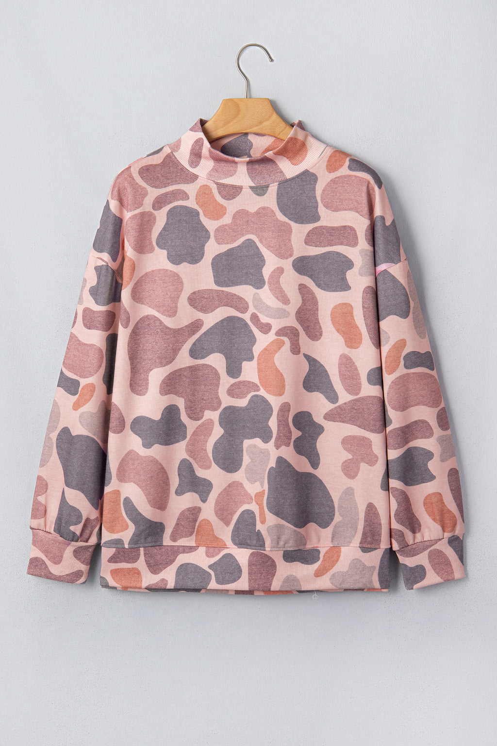 The Camo Mock Neck Pullover Sweatshirt features long sleeves, a high neckline, and a relaxed tunic silhouette in a pink and mauve camouflage print. It is displayed on a wooden hanger against a light gray wall.