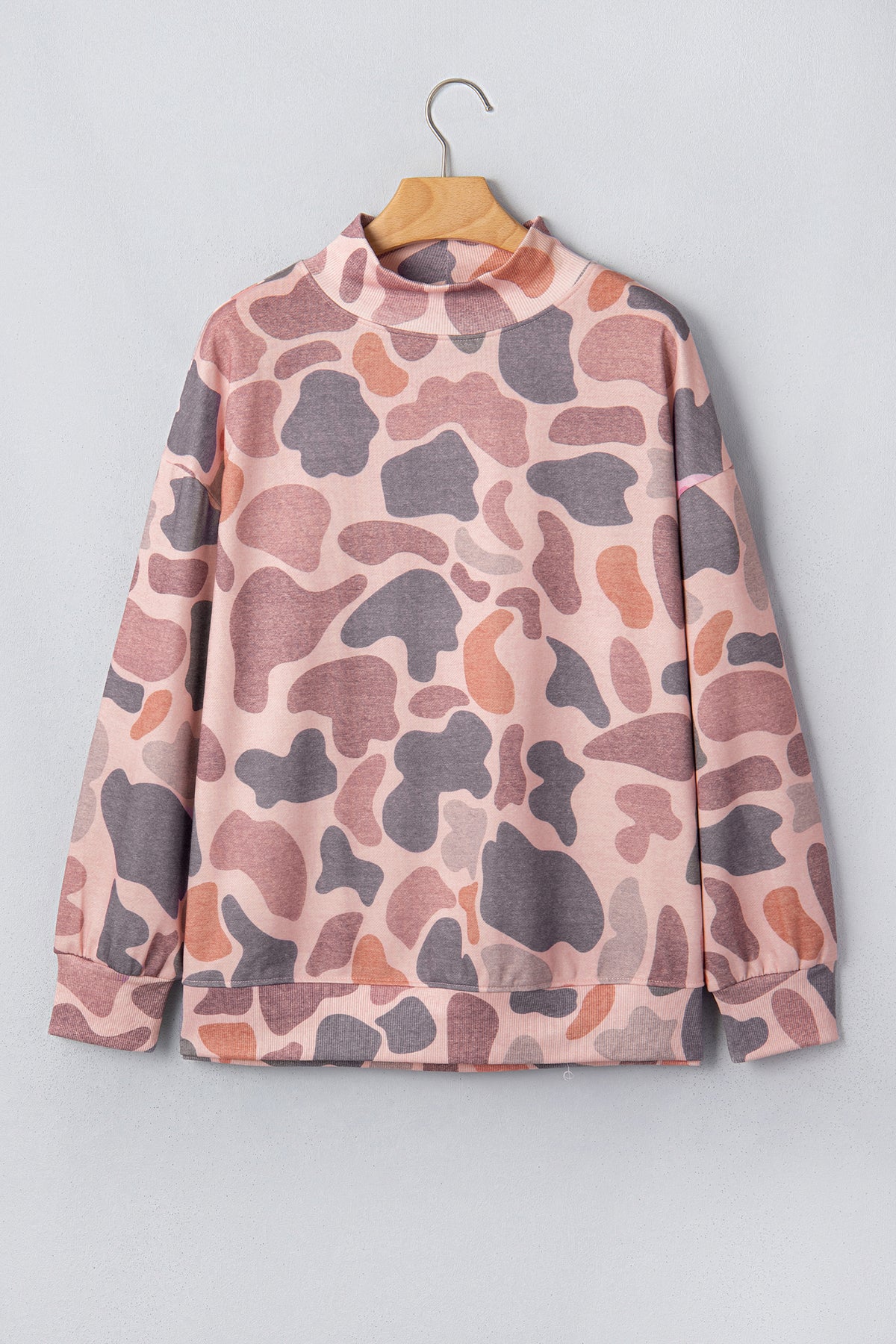 The Camo Mock Neck Pullover Sweatshirt features long sleeves, a high neckline, and a relaxed tunic silhouette in a pink and mauve camouflage print. It is displayed on a wooden hanger against a light gray wall.