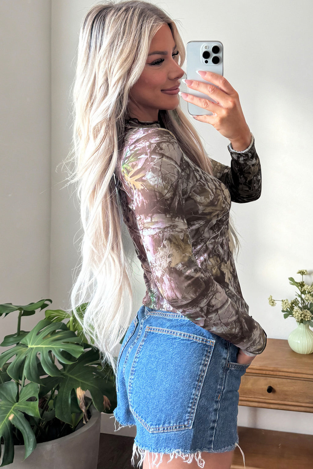 A woman with long blonde hair takes a mirror selfie, wearing the Smoke Gray Floral Mesh Mock Neck Top and denim shorts. There are green plants and a small vase of flowers in the background.