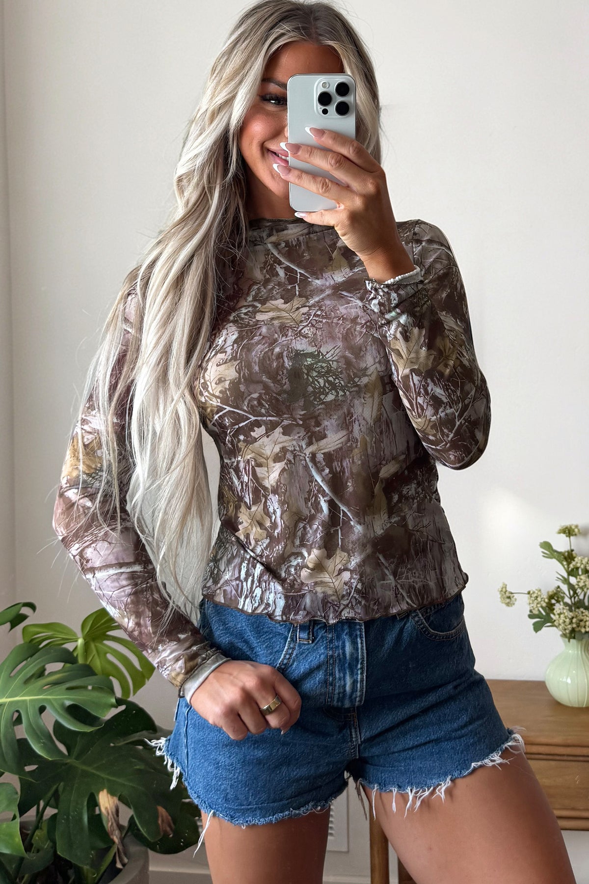 A woman with long, wavy blonde hair takes a mirror selfie. She wears the Smoke Gray Floral Mesh Mock Neck Top, which features a slim silhouette and long sleeves, paired with frayed denim shorts. Plants and a small vase with flowers are visible in the background.